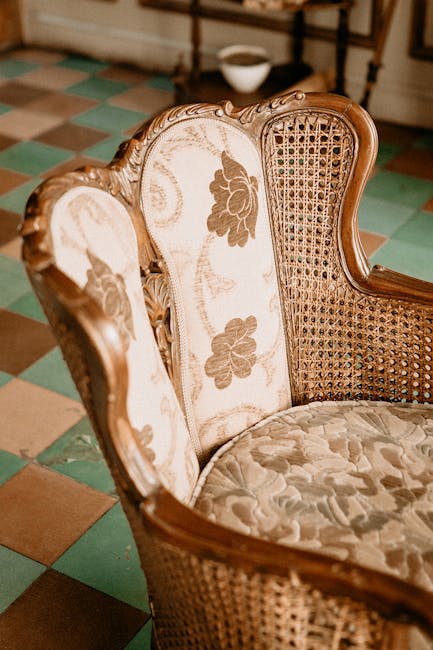 A close-up view of an antique-style upholstered armchair with a wooden frame, featuring floral-patterned fabric on the cushions and woven cane detailing on the backrest, situated on a checkered tile floor with green and beige squares. The room has soft, natural lighting, highlighting the fabric's cleanliness and the wood's polished finish, indicative of professional deep cleaning and surface sanitisation. The background shows a portion of a rustic wall and a white bowl placed nearby, suggesting a period home's interior setting. This image exemplifies domestic cleaning and maintenance of period furniture, aligning with services offered by Carpet Cleaning Holland Park for enhancing hygiene and preserving the appearance of historic home furnishings.