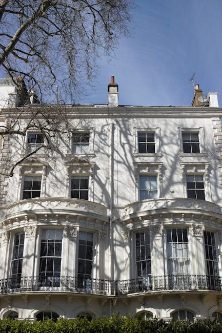 A historic white Victorian terraced building with detailed architectural features including bay windows, decorative cornices, and iron balcony railings, located on Pembridge Villas in Holland Park, under a bright blue sky. Shadows of leafless tree branches are cast on the facade, and the windows are clean and well-maintained. The scene suggests a residential setting with a well-preserved exterior, characteristic of period homes. Carpet Cleaning Holland Park specializes in interior and upholstery cleaning for such traditional homes, providing comprehensive deep cleaning and sanitisation services to uphold the property's aesthetic and hygiene standards.